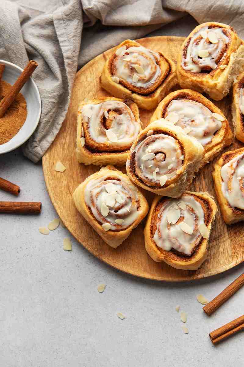 Eine Draufsicht zeigt ein rundes Holzbrett belegt mit mehreren köstlichen Zimtschnecken. Die charakteristischen Zimtwirbel sind gut sichtbar, und die Schnecken sind mit einem weißen Zuckerguss überzogen und mit gehobelten Mandeln bestreut. Einige lose Mandelsplitter und Zimtstangen liegen dekorativ um die Zimtschnecken auf einer hellgrauen Oberfläche. Oben rechts sind ein gefaltetes graues Stoffserviette und eine kleine weiße Schale angeschnitten.